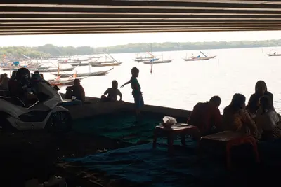 People sitting in shade under a bridge by the water, a boy standing on the wall, fishing boats moored beyond.