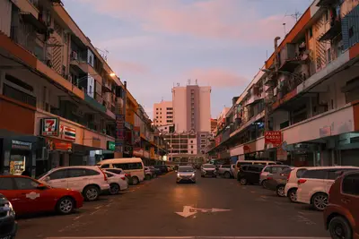 A street of low shophouses at dusk under a pink sky, cars parked along both sides.