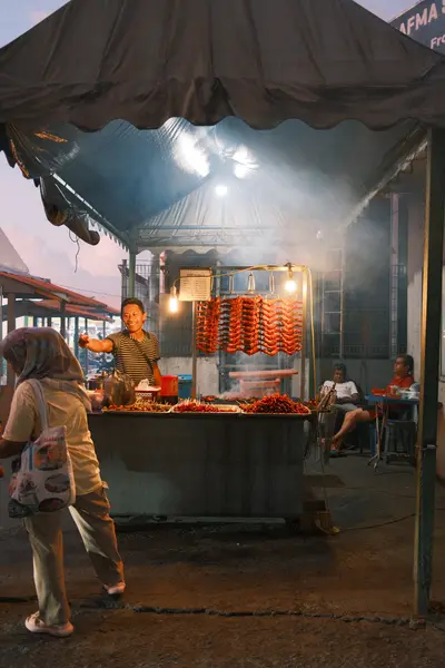 A street food stall at dusk, smoke rising from the grill under a canopy while a woman waits.