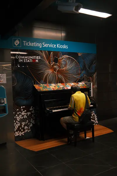 A man in a yellow shirt playing a painted public piano in a dark train station.